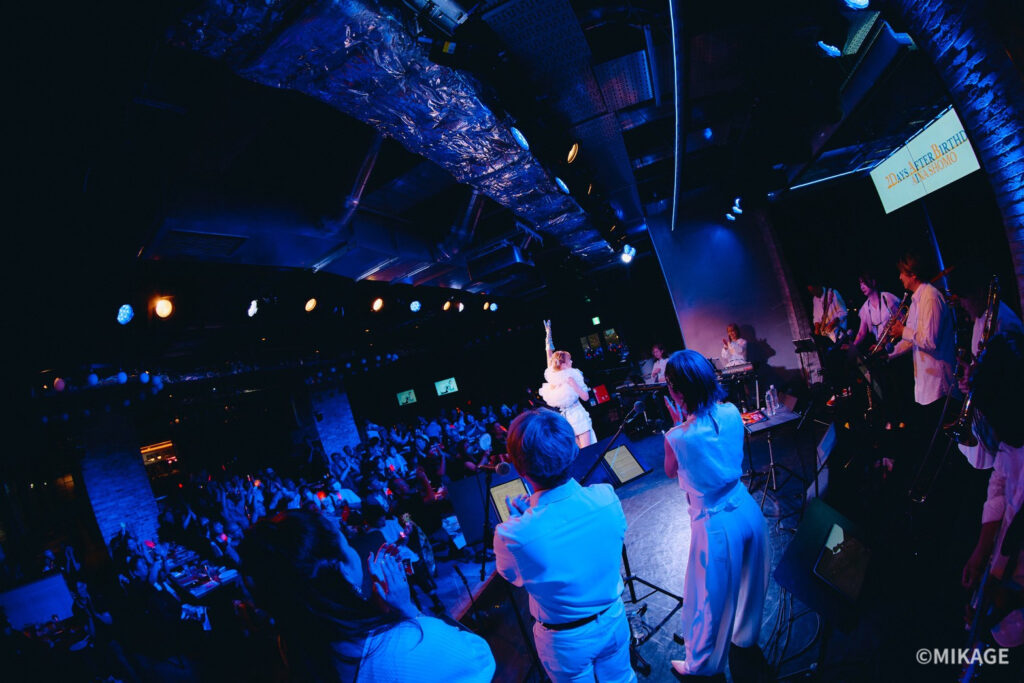Band performing on stage under blue lighting, singer with arm raised, crowd cheering in a venue