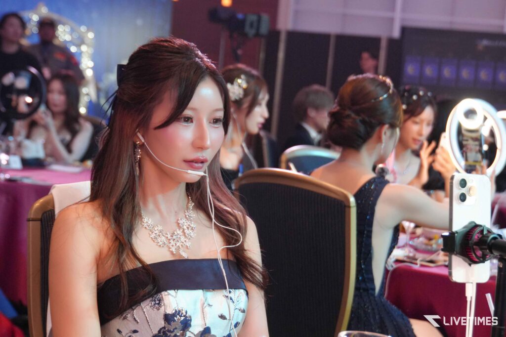 Close-up of a woman wearing a headset mic at a booth with a ring light and other attendees blurred in the background.