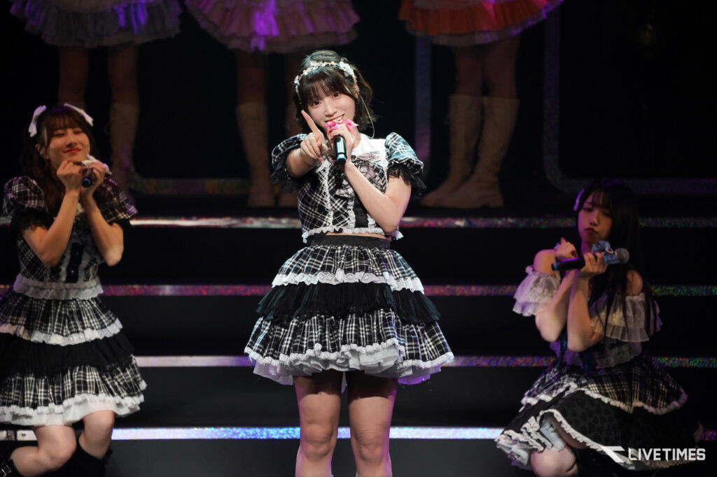 Center-stage young singer in a black-and-white plaid outfit points to the audience while singing into a microphone, flanked by two dancers on a dark stage.