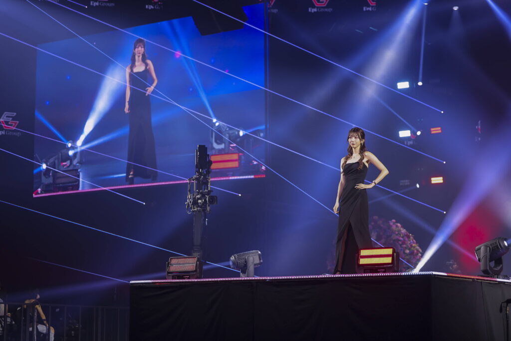 Two women in black evening gowns pose on a brightly lit stage as blue lasers streak across the air during a live performance, with a camera rig in the foreground.