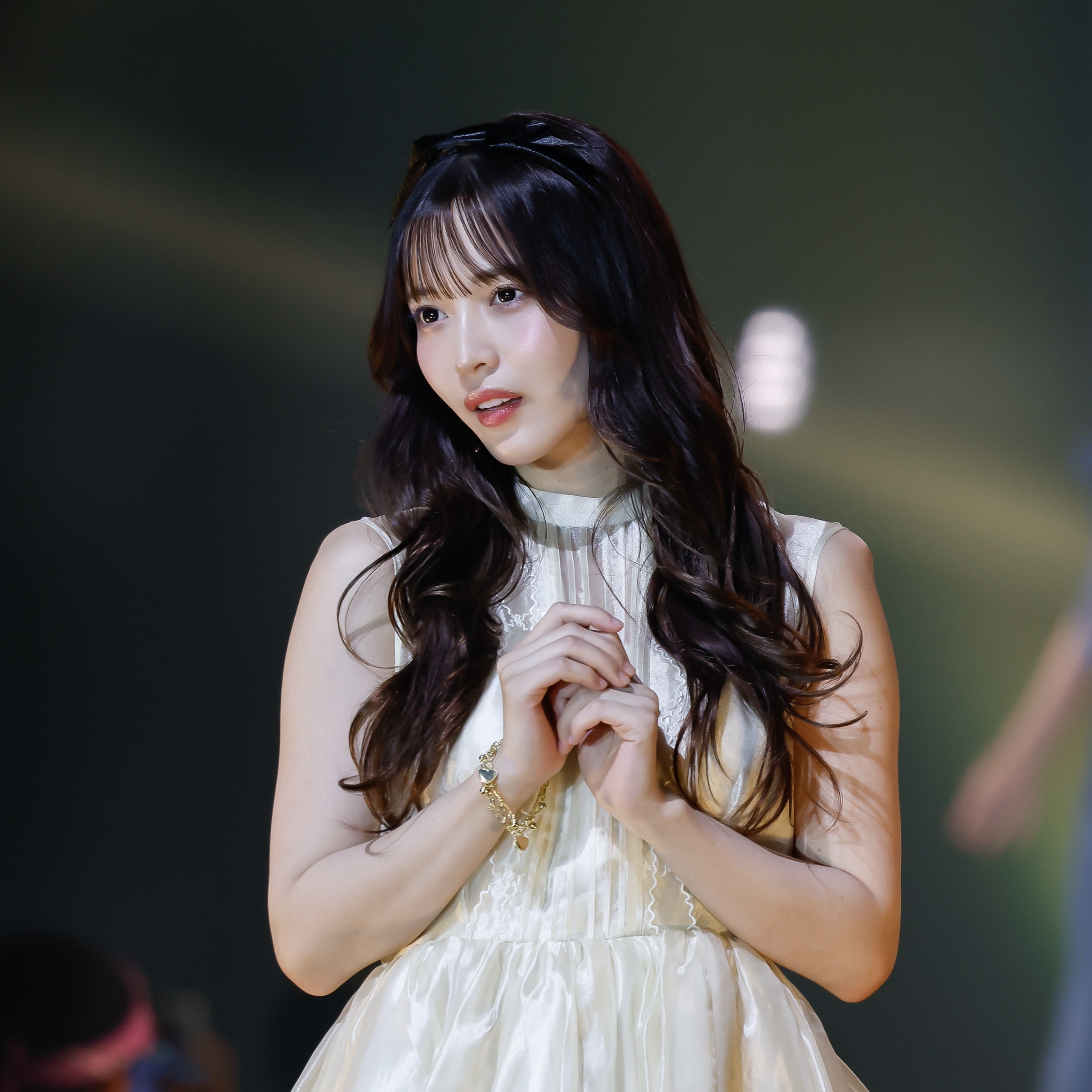 Young woman in a white dress on stage, hands clasped near her chest, looking slightly to the side.