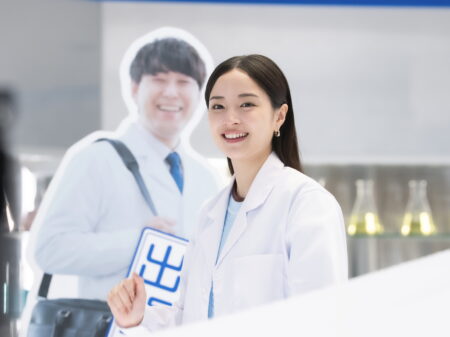 Smiling woman in a white lab coat holding a large ID badge; a man in a cutout poster stands behind her.