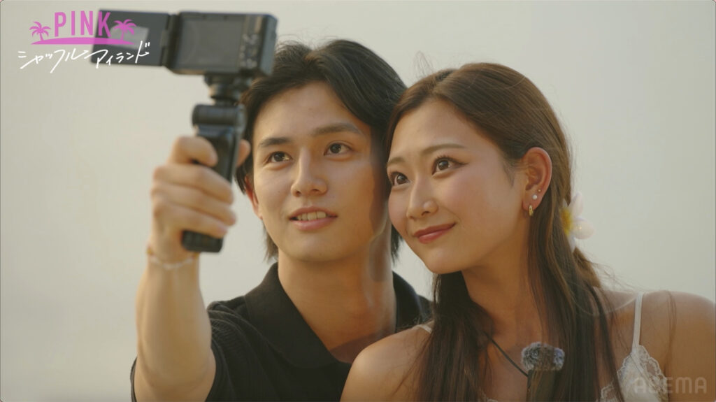Two young adults pose for a selfie with a handheld camera, logo 'PINK' and palm trees in the top-left corner.