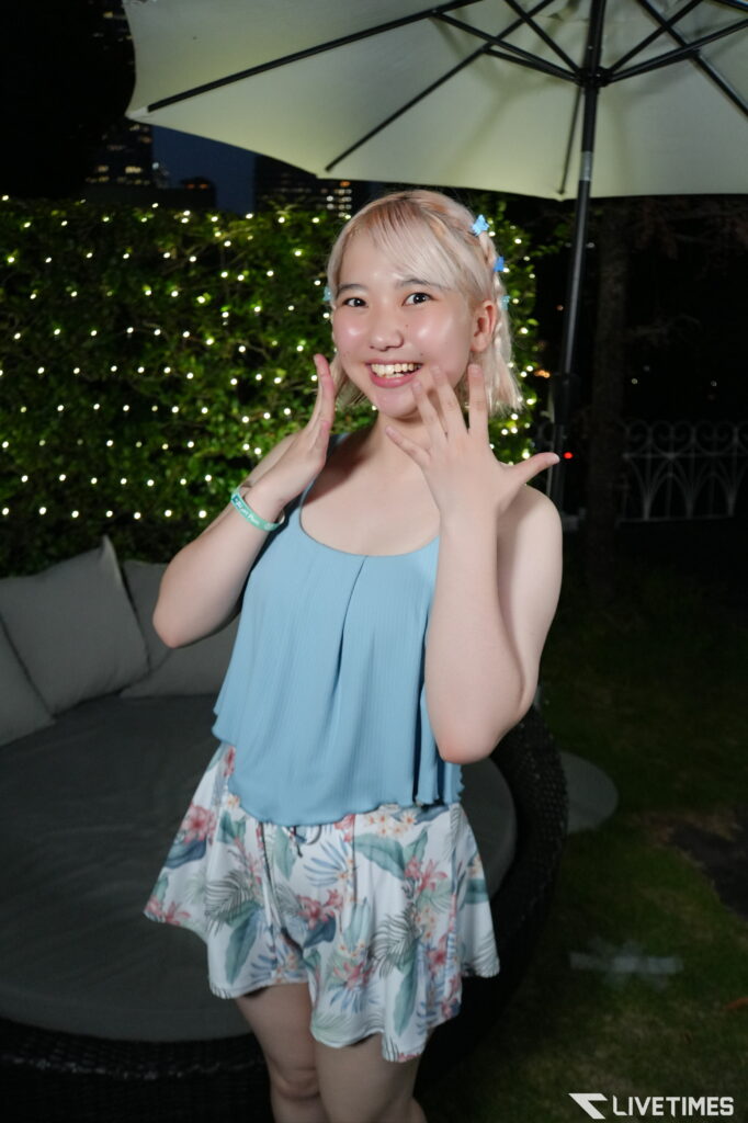 Smiling girl in a light blue top and floral skirt posing with hands near her face during a nighttime outdoor gathering with string lights behind her.