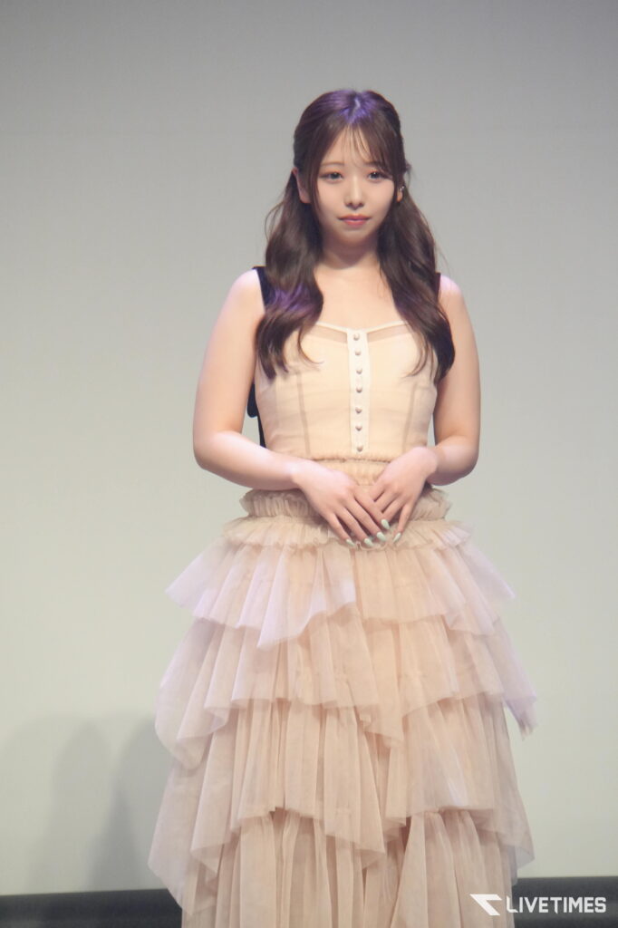 Young woman on stage wearing a pale pink, layered dress with hands clasped in front of her.