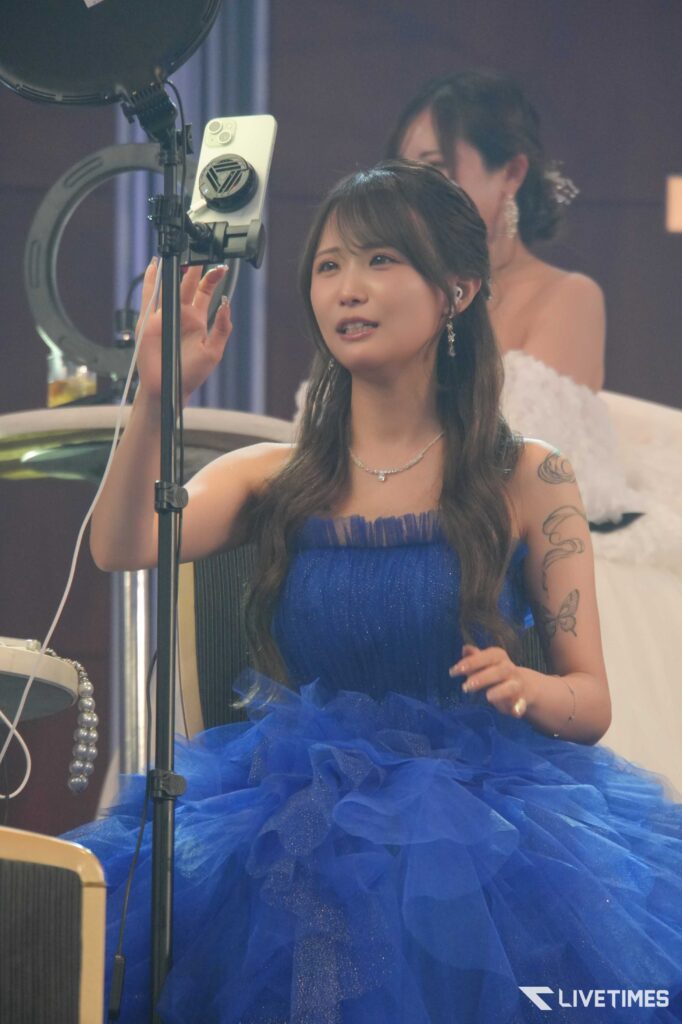 Female singer in a blue, ruffled gown raises her hand toward a microphone on a stand during a performance on stage with studio lighting.