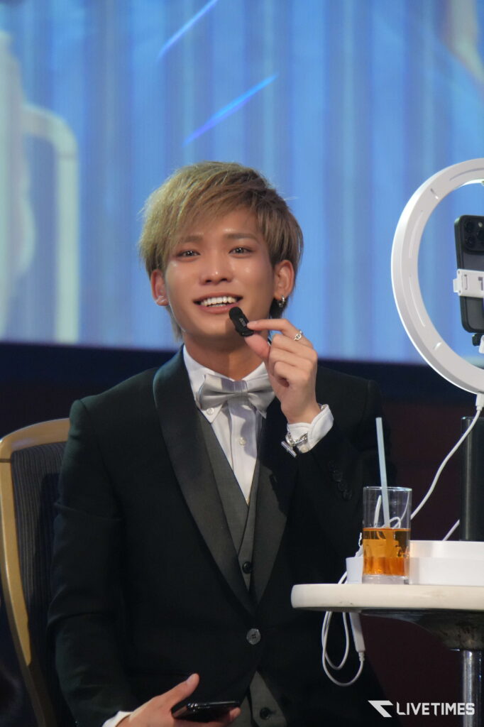 Young man in a black tuxedo and bow tie, smiling, holding a small black device near his mouth on a stage with blue backdrop.