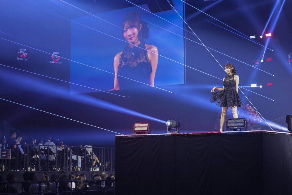 A female singer in a black lace dress performs on a blue-lit stage with laser lines and a large screen behind her.
