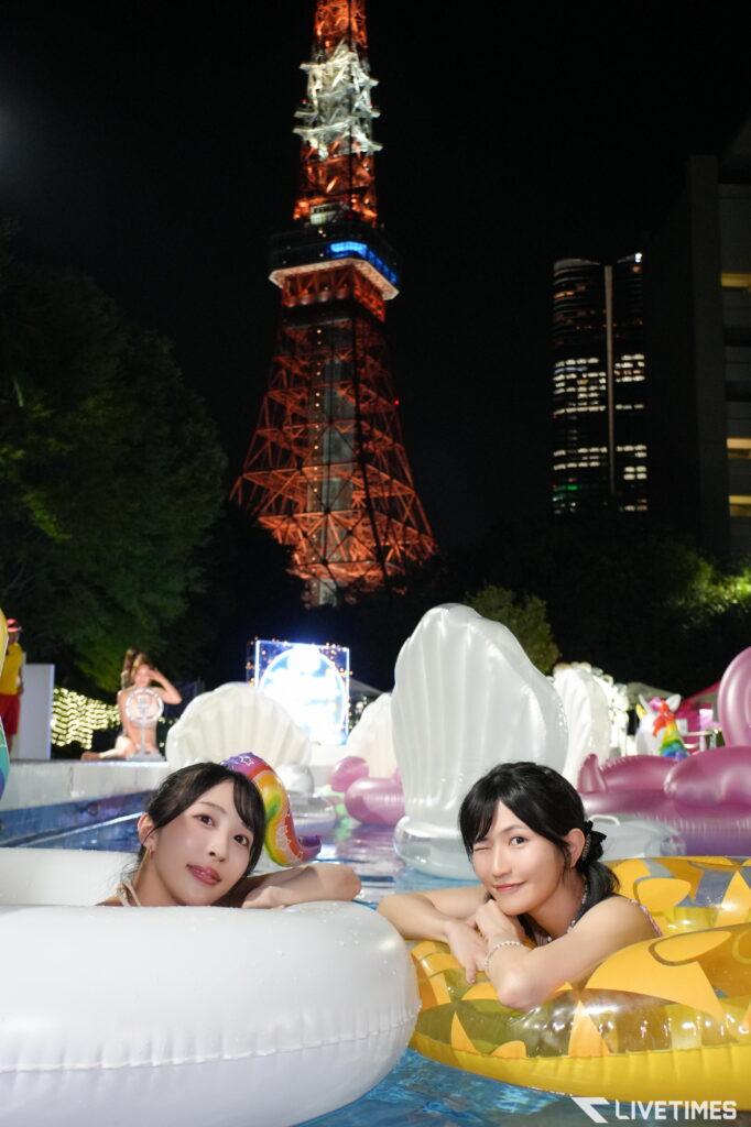 Two women relax in a pool on inflatable rings at night with a lit tower in the background, city lights around them.