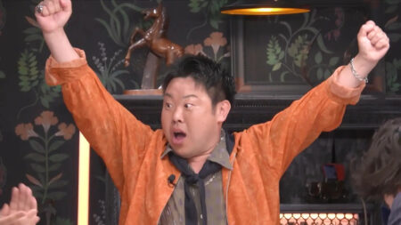 Man in an orange jacket raises both arms in celebration, mouth open in a shout, in a floral, dimly lit setting.