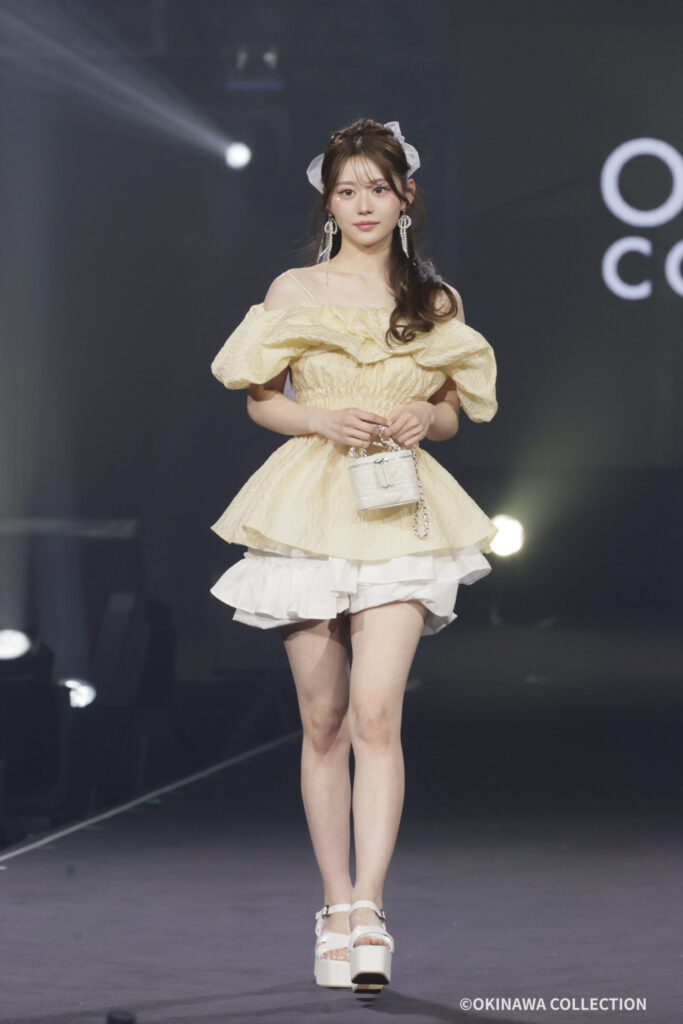 Model walks the runway in a pale yellow off-shoulder dress with puff sleeves, white layered skirt, platform sandals, and a small white handbag.