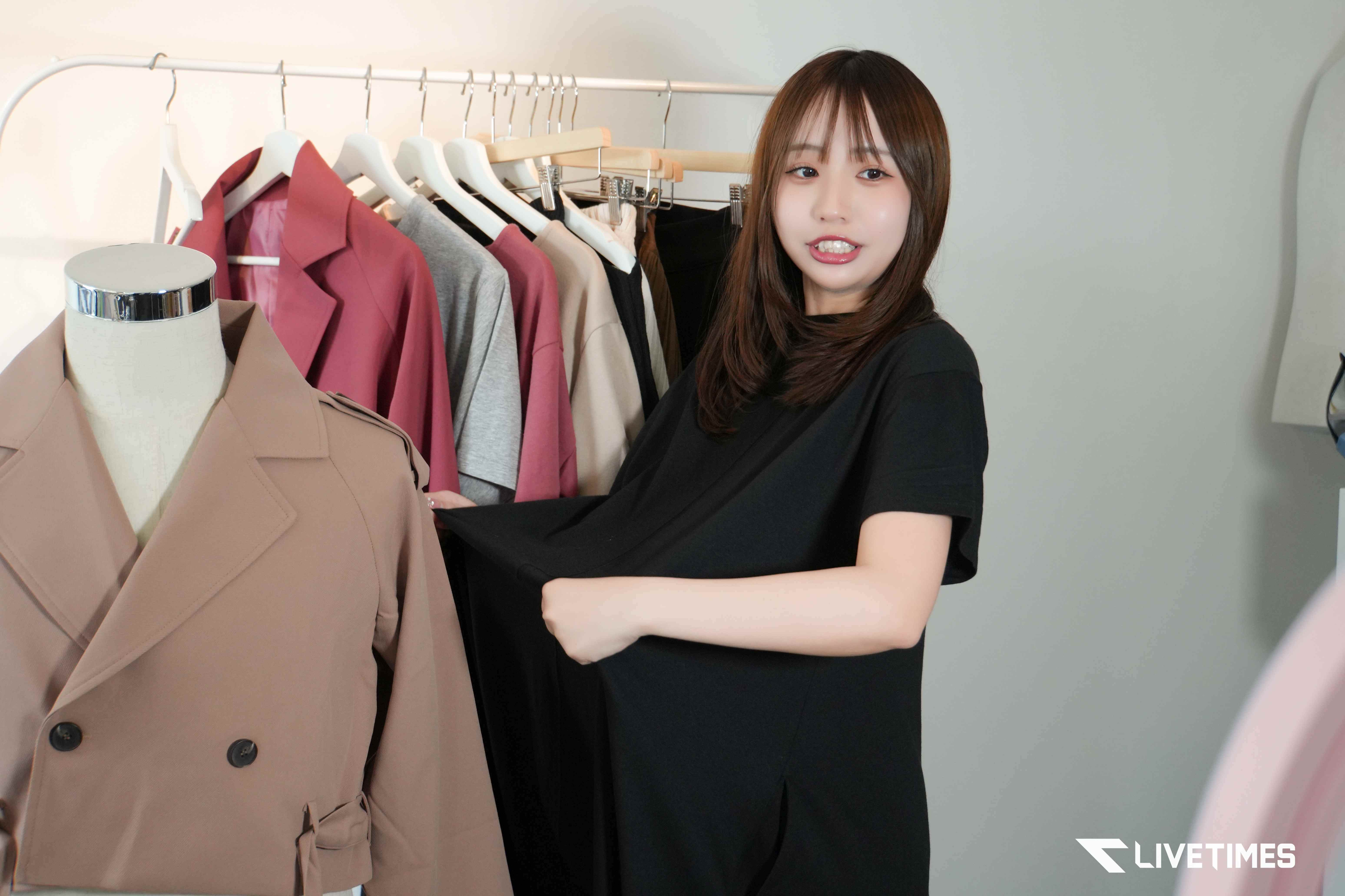 Woman in a black top browses a rack of garments; beige trench coat on a mannequin sits in front.