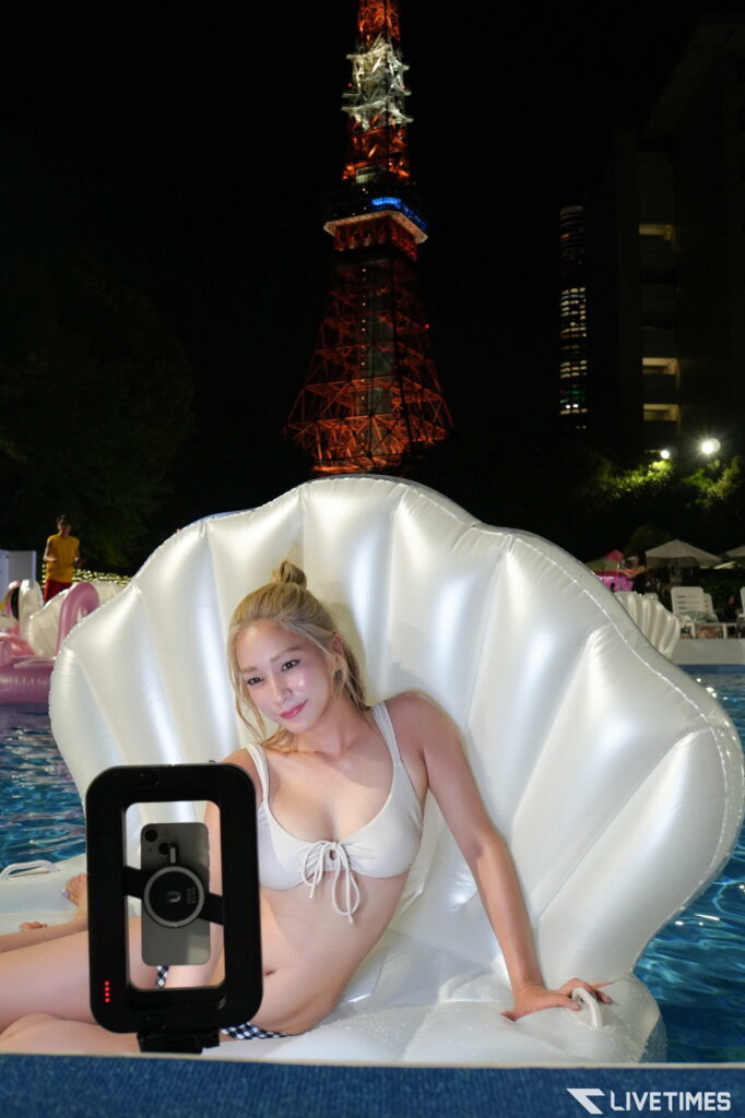 Woman in a white swimsuit sitting in a large white seashell float, taking a selfie with a mounted phone in a nighttime pool; a lit tower rises in the background.