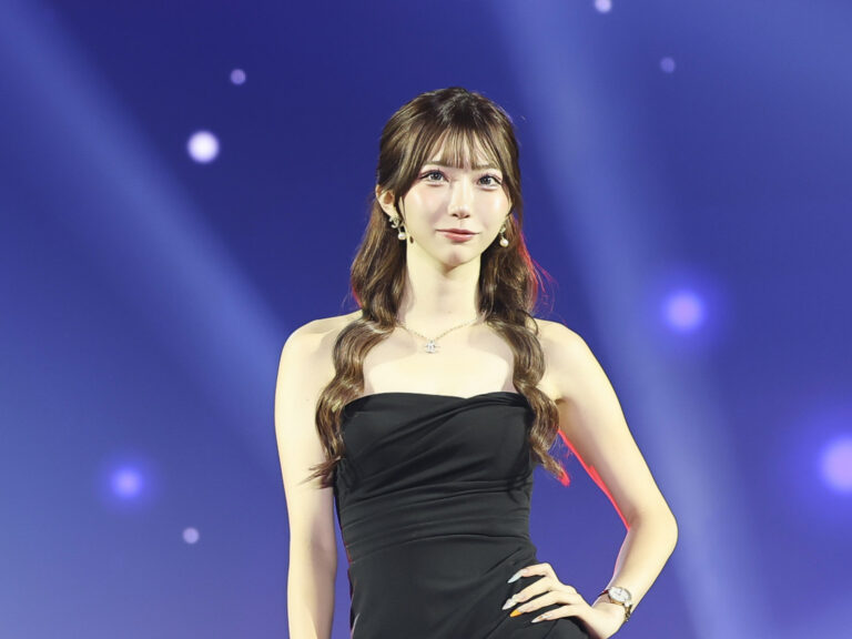 Young woman in a black strapless dress posing with hand on hip on a stage with blue spotlight background while wearing jewelry.