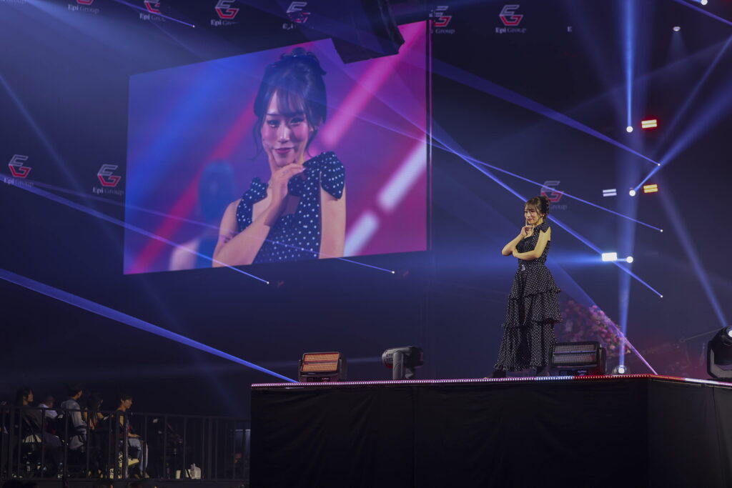 Singer on stage with a large screen showing her close-up, wearing a black polka-dot dress amid colorful lights.