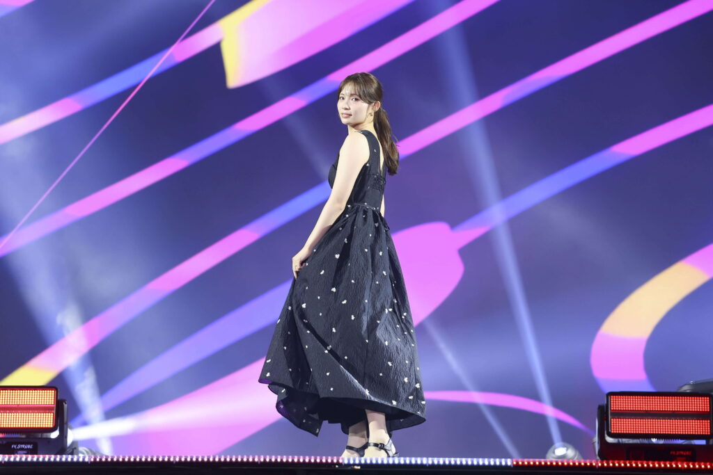 Female performer on stage in a dark dress, looking over her shoulder as colorful neon lights streak behind her.