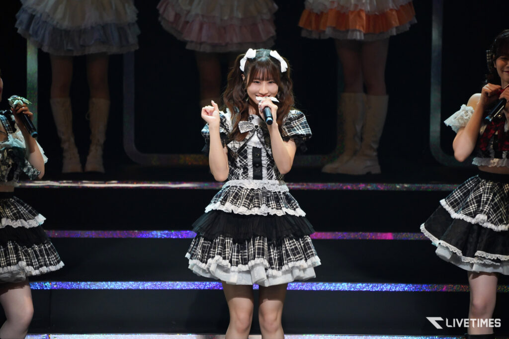 Center-stage female singer in a black-and-white plaid dress with lace trim, holding a microphone and smiling; backup dancers in matching outfits on a dark stage behind her