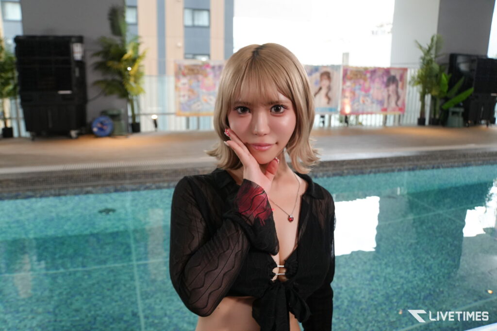 Young woman with blonde bob posing by a pool, hand resting on her cheek, wearing a sheer black top with a red pendant necklace.
