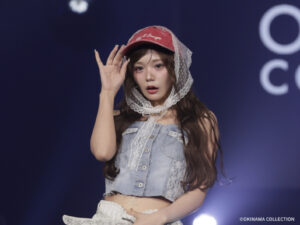 Young performer on stage wearing a lace veil and red cap, waving to the audience