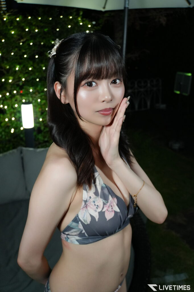 Young woman in a floral bikini top posing with hands near her face at night, decorative lights in the background.