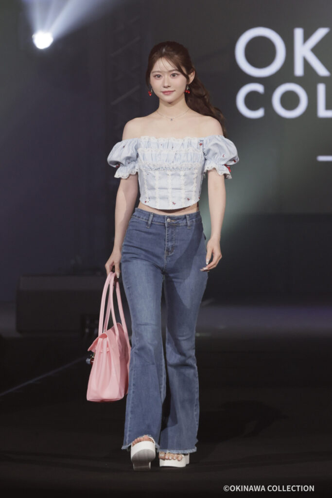 Model walking a fashion runway in an off-shoulder blue striped top, high-waisted jeans, white platform sandals, and a pink handbag; red earrings add.