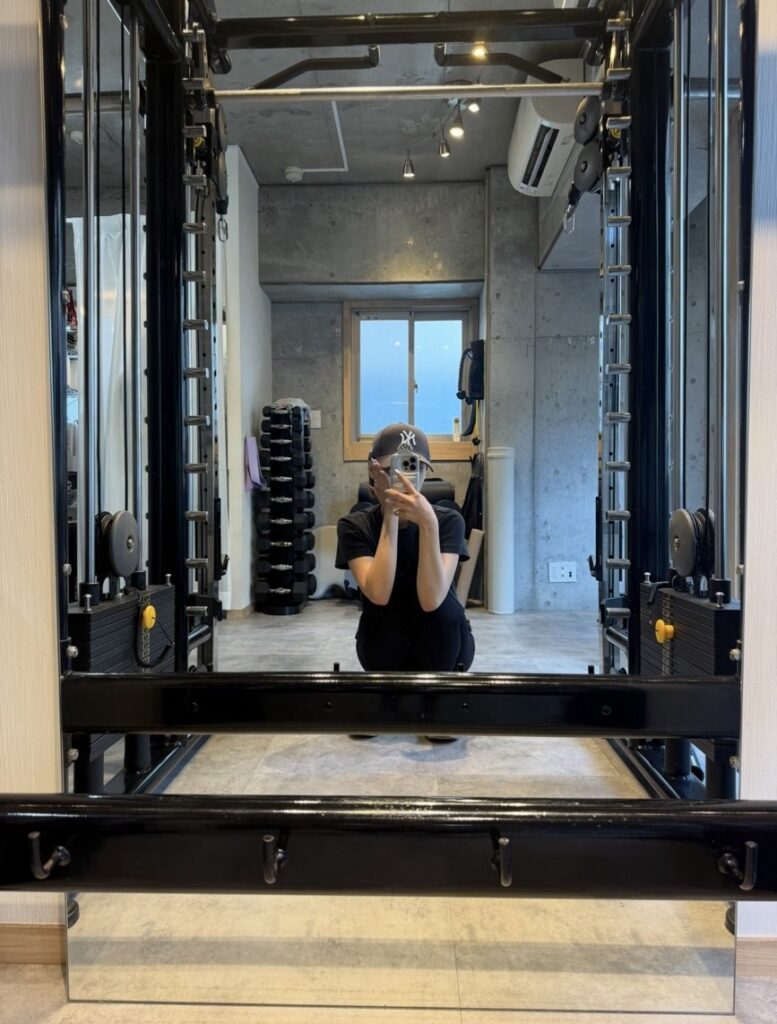 Person in a black cap and shirt crouches in a gym squat rack, taking a mirror selfie with a smartphone in front of them.