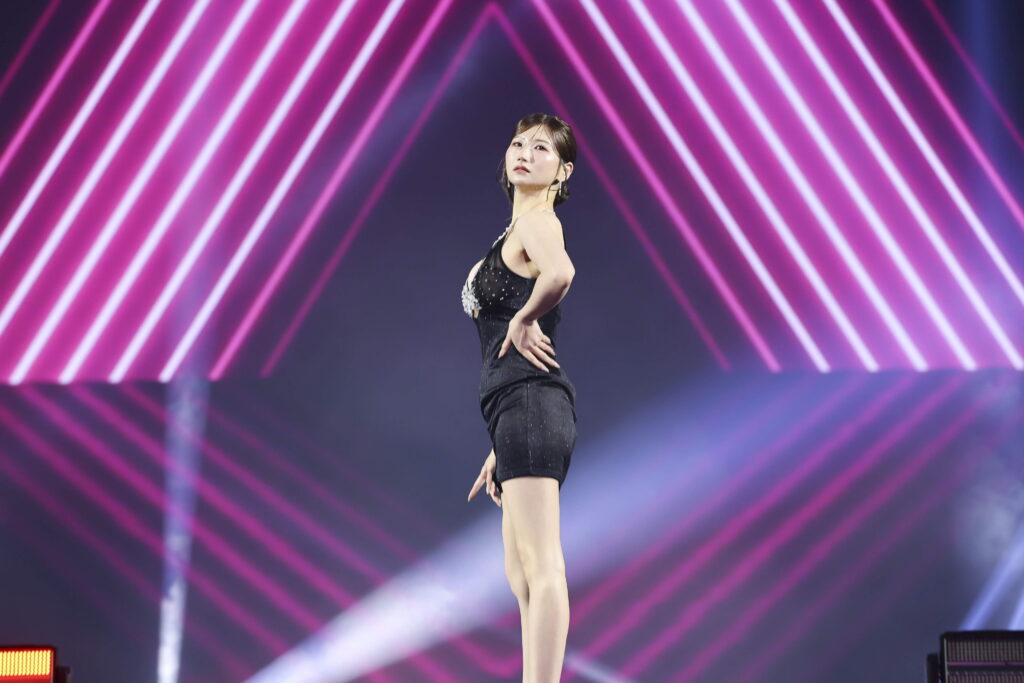 Female performer in a glittery black dress posing with hand on hip on stage, neon pink diagonal lights in the background.
