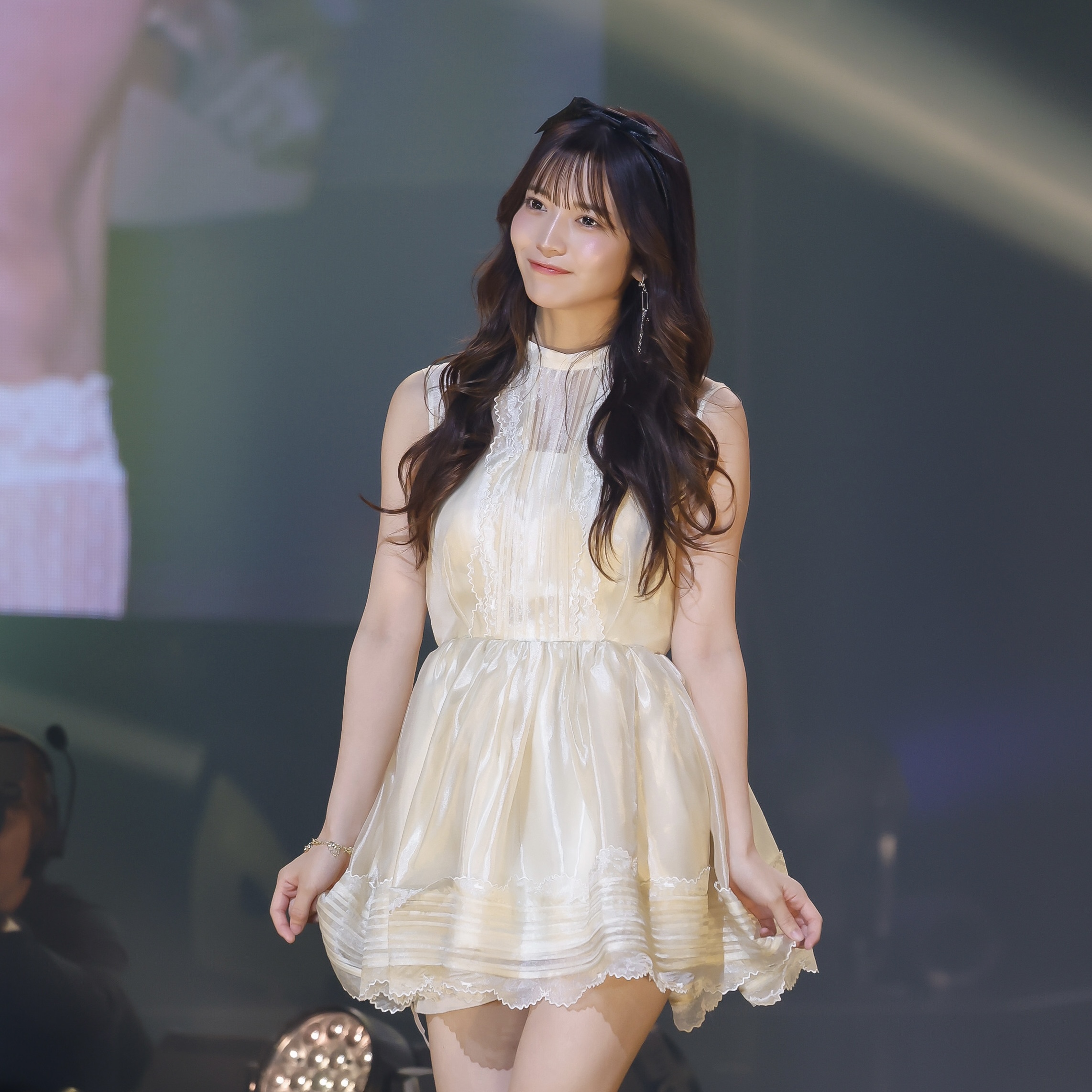 Young woman on stage wearing a ivory lace dress, looking slightly to the left with a soft smile.