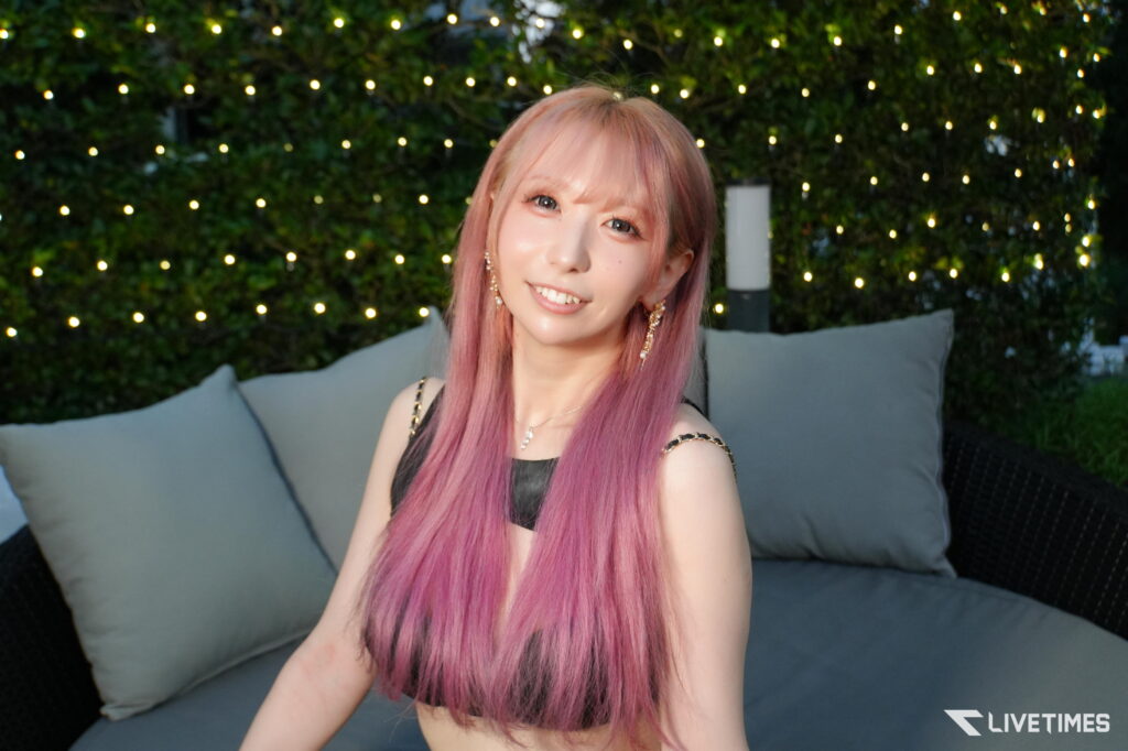 Portrait of a smiling woman with long pink hair and jewelry, seated on a sofa with twinkling string lights in the background.