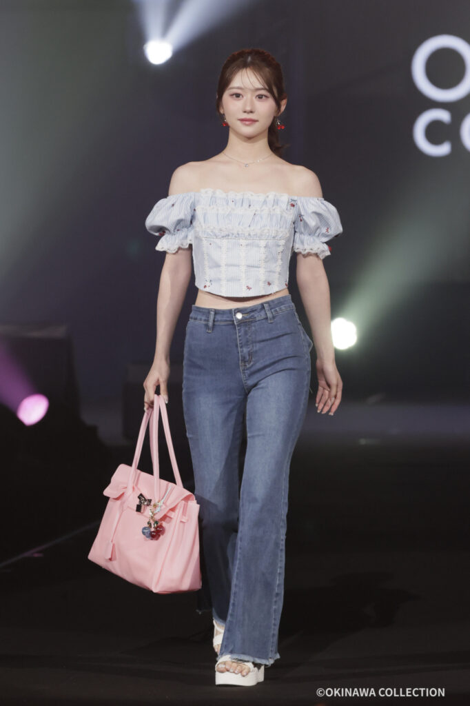 Model walking a fashion runway in an off-shoulder blue striped top and high-waisted jeans, carrying a pink handbag under bright stage lights.