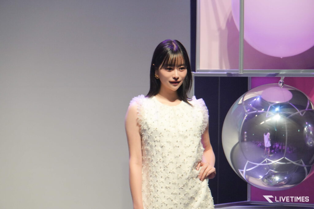 Young woman in a white beaded dress standing on a stage beside a large purple-glowing glass sphere display.