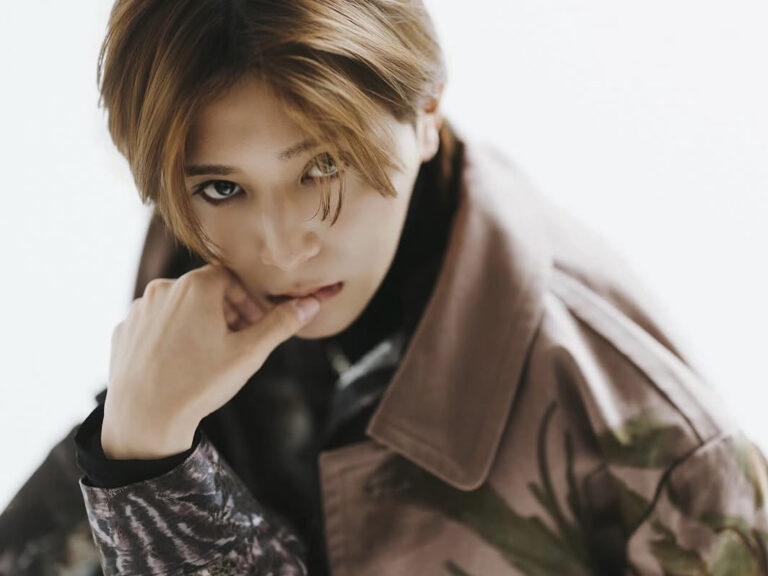 Young person with short blond hair wearing a camo jacket, touching lips with their hand and looking at the camera.