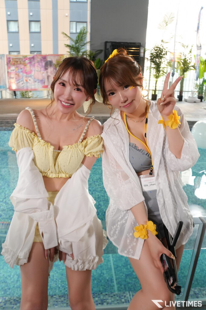 Two smiling young women in yellow and white outfits posing by a pool, one flashing a peace sign against a backdrop of plants and a building.