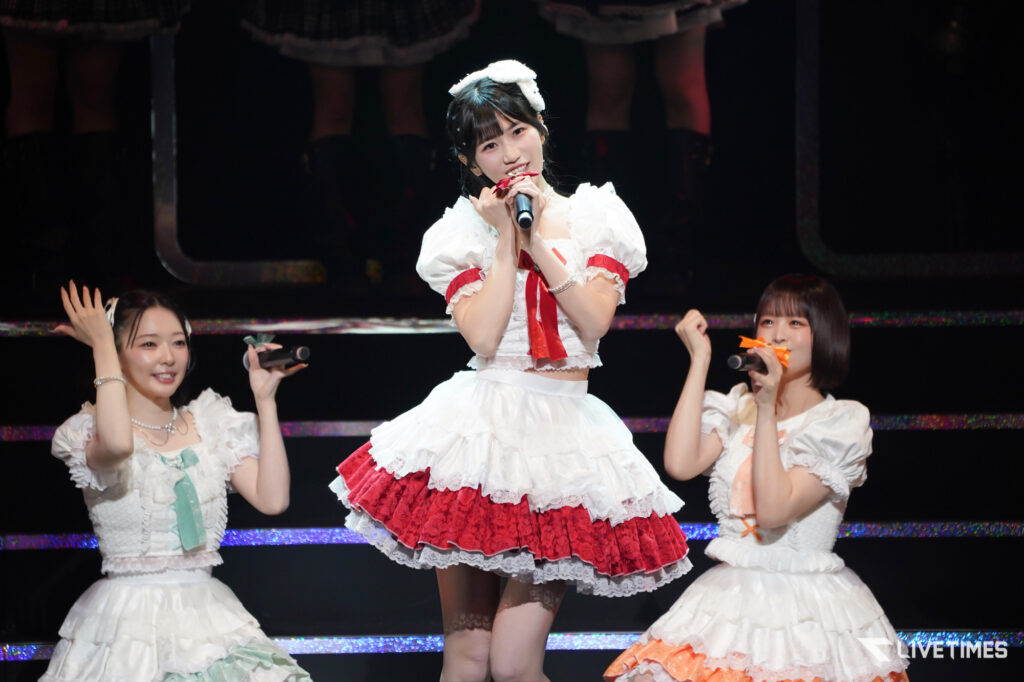 Center-stage singer in a white and red frilly dress, holding a microphone, flanked by two dancers in white outfits on a lit stage
