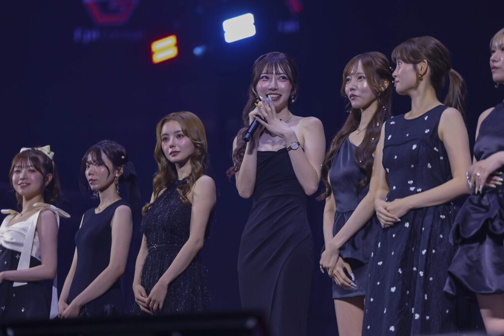 Group of female performers in elegant navy outfits on stage, central woman speaking into a microphone while other members stand nearby.