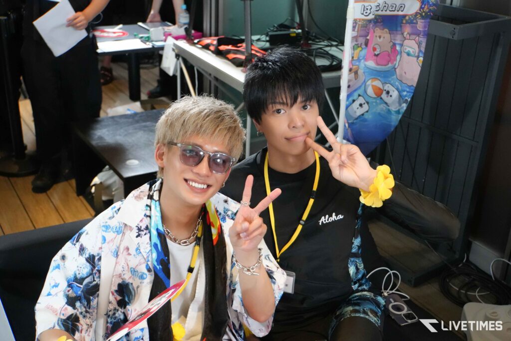 Two young men pose for a selfie, flashing peace signs at a crowded indoor event; one in a floral shirt, the other in black with a yellow lanyard and a yellow flower bracelet visible on his wrist behind them.