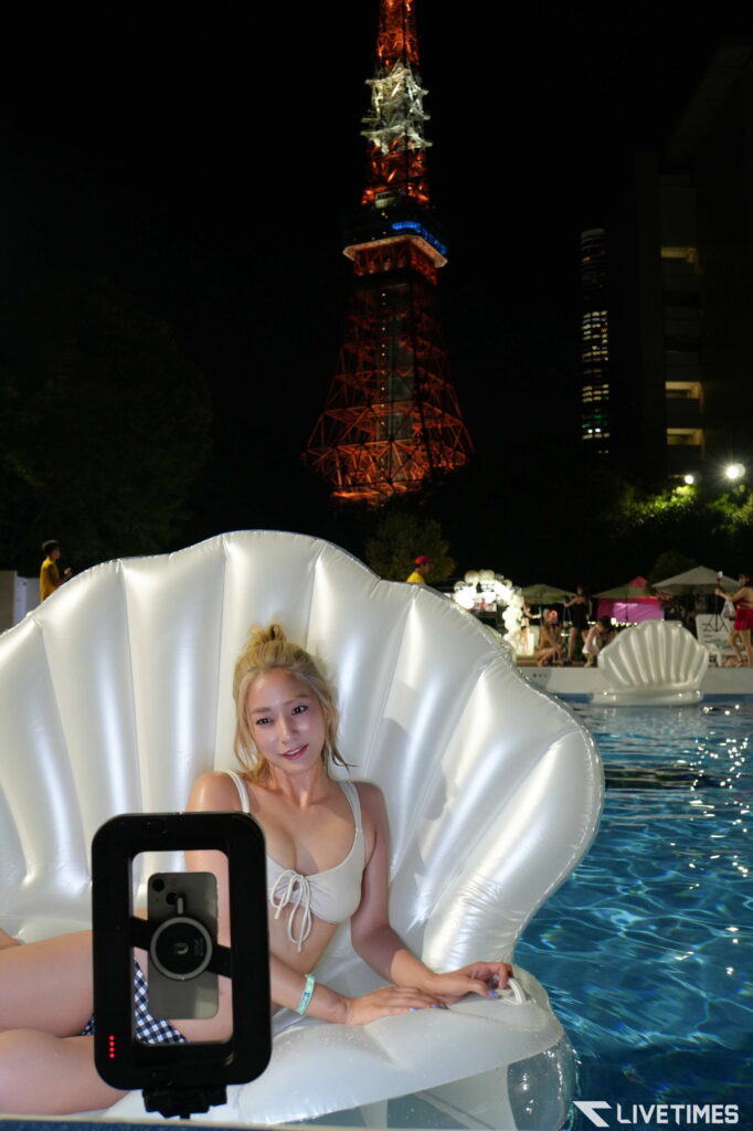 Woman in a white swimsuit sits in a large white pool float at night, taking a selfie with a phone mounted on a stand; Tokyo Tower lights shine in the background.