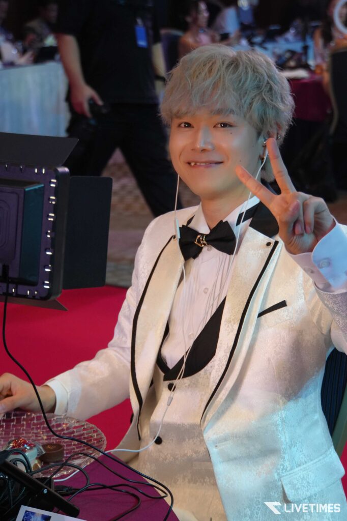 Smiling young man in a white tuxedo making a peace sign at a desk with cables and equipment, event background.