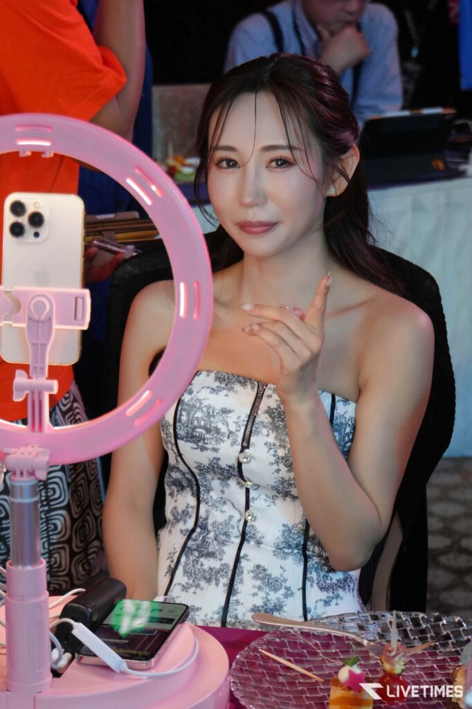 Woman posing for a selfie at a pink ring light with a smartphone nearby for streaming or filming.