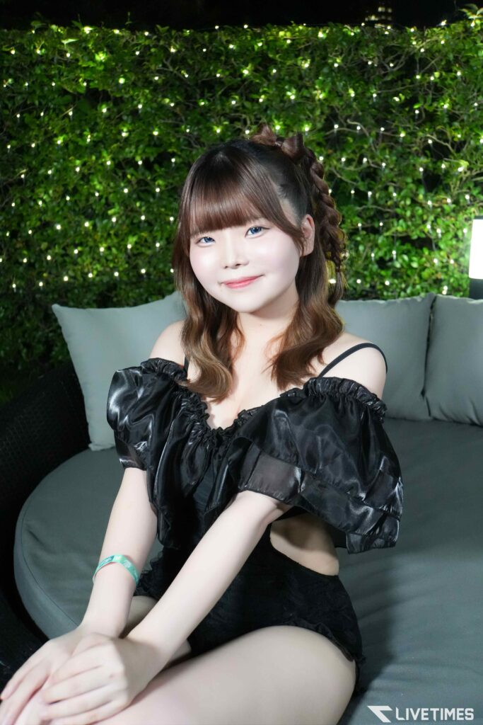 Smiling woman with brown hair in a black off-the-shoulder dress seated on a gray sofa, green hedge and twinkling lights behind her.