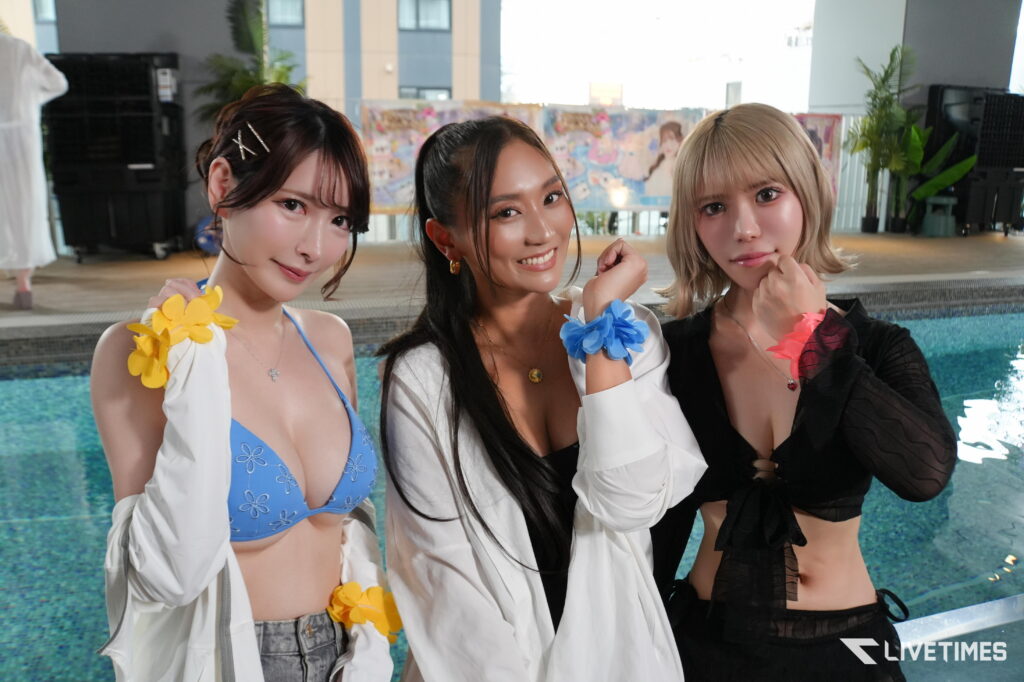 Three young women pose by a pool, wearing bikinis and flower accessories, smiling at the camera.