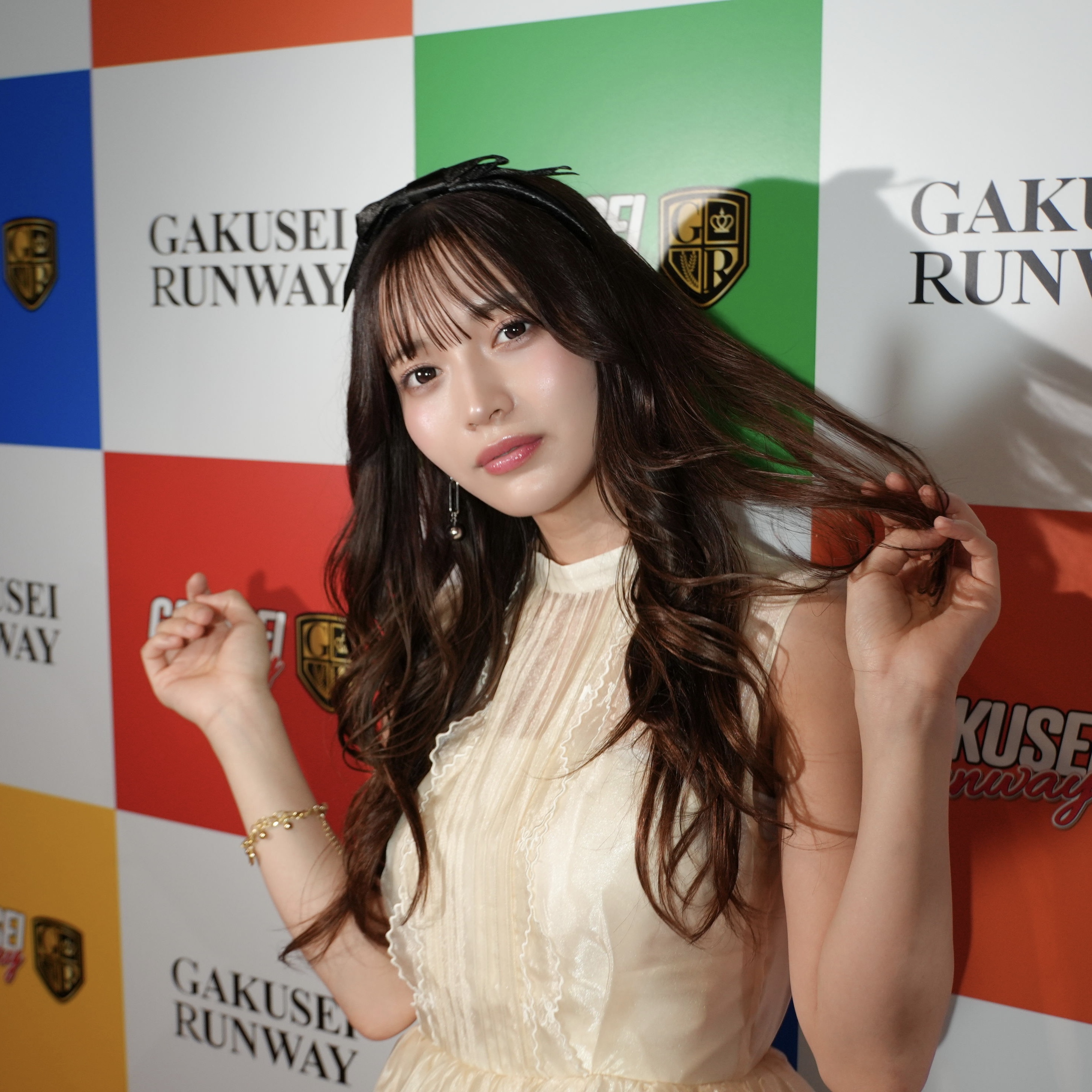 Young woman with long dark hair in a cream dress posing in front of a colorful event backdrop with logos and shields.