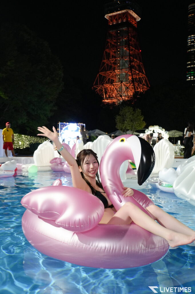 Woman relaxing in a pink flamingo inflatable pool float at night with a red-lit tower in the background and party lights nearby