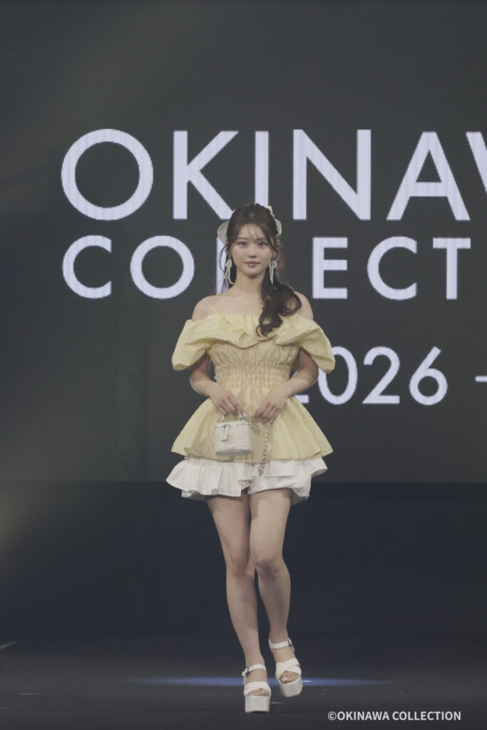 Model in pale yellow off-shoulder dress on a runway with large 'OKINAWA COLLECTION' backdrop, holding a small white bag.