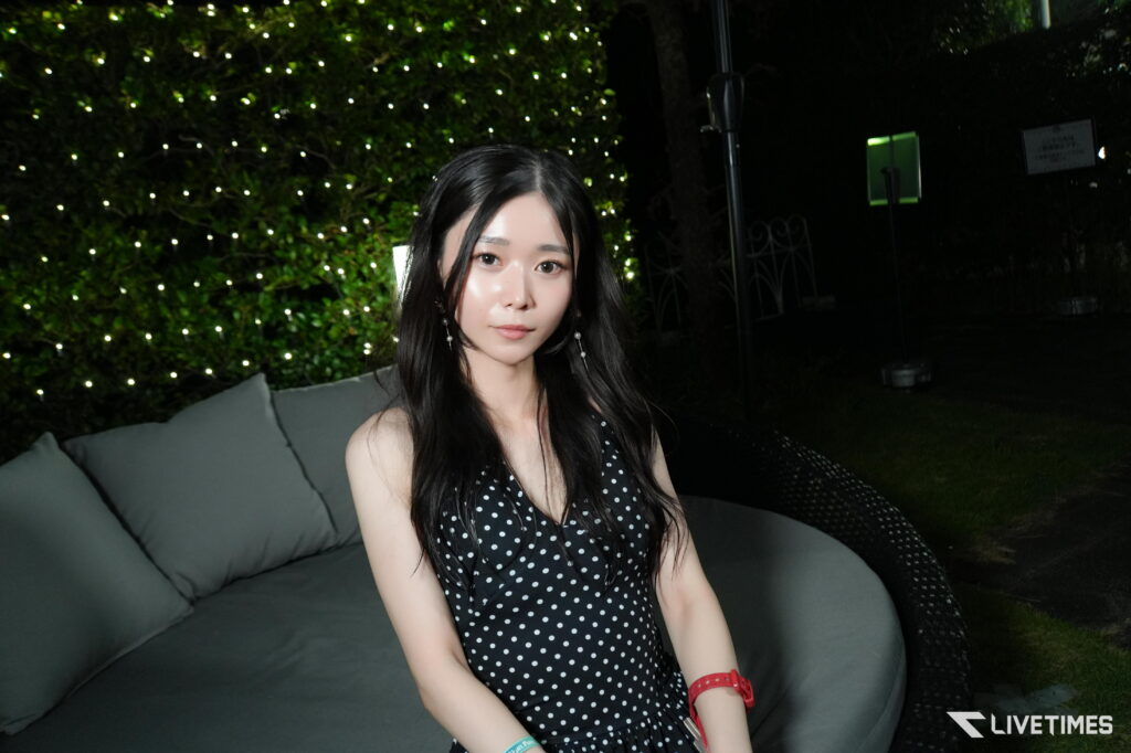 Woman with long dark hair in a black polka-dot dress sits on a gray outdoor sofa at night, illuminated by background fairy lights.