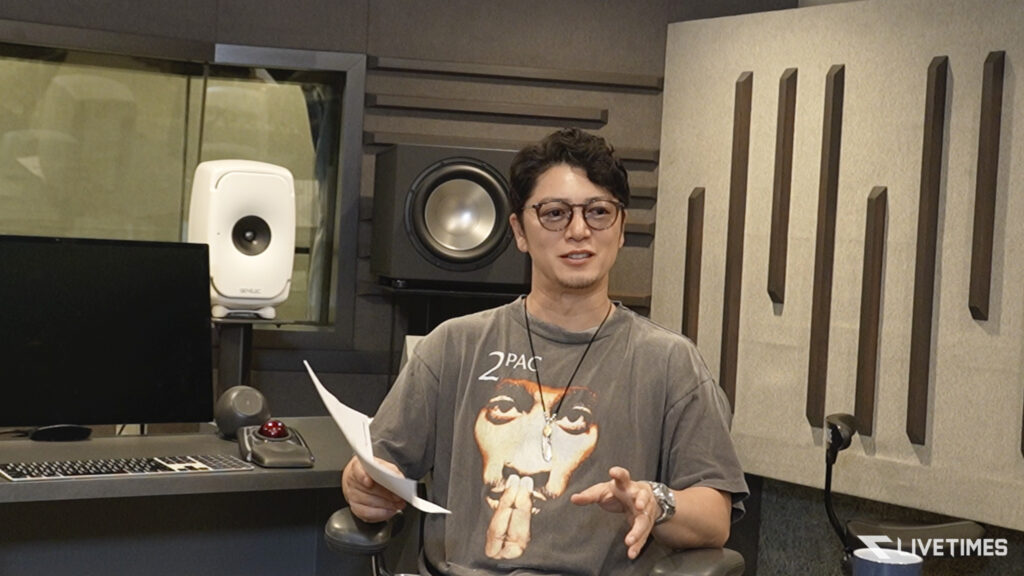 Man with short dark hair and glasses speaks while holding papers in a recording studio, with studio monitor, keyboard, and acoustic panels around him.