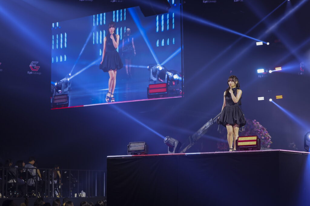 Two female performers in black dresses on a lit concert stage with blue spotlights and a large video screen behind them.
