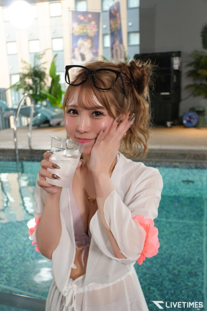 Woman by an indoor pool holding a glass of water, sunglasses perched on her head, wearing a white outfit with pink accents.