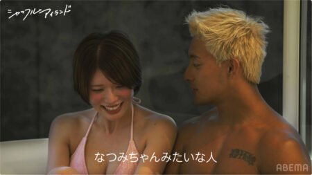 Smiling woman in a pink bikini sits beside a shirtless man with bleached blond hair in a hot tub, with white Japanese caption overlaid at the bottom.