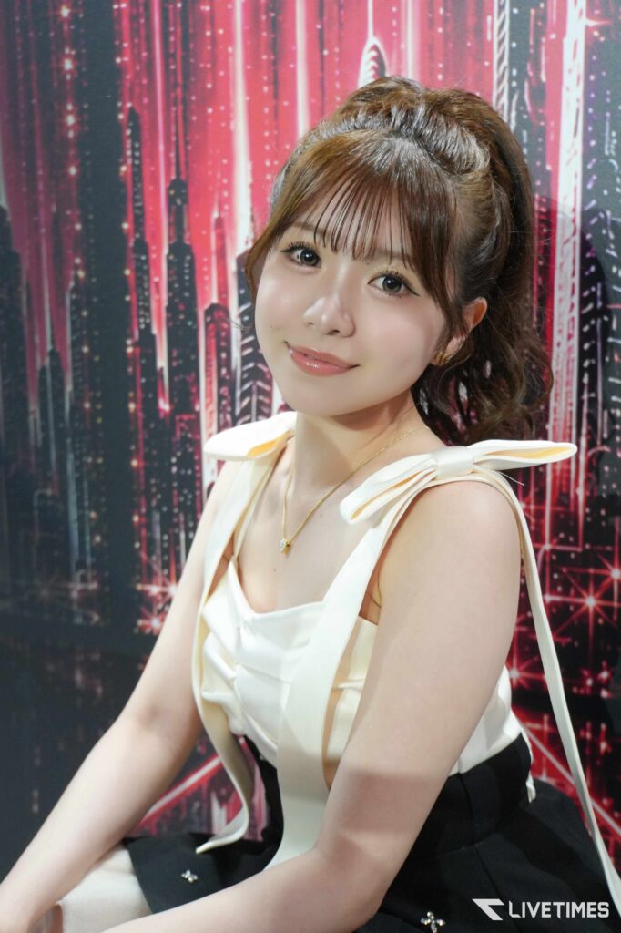 Young woman with brown hair in a high ponytail, wearing a cream sleeveless top, posing against a red-pink stellated backdrop.